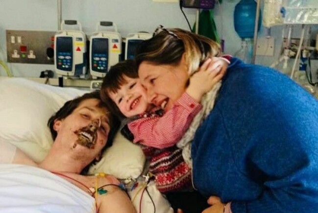 Alex Lewis ligger i en sjukhussäng och bredvid står frun Lucy med sonen Sam i famnen.