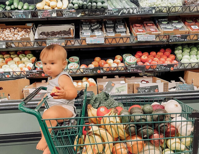 Tvååriga Fern äter bara ekologisk, naturlig och vegansk kost. Fern sitter i en kundvagn fylld med grönsaker och frukt.