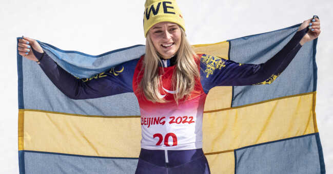 Ebba Årsjö har vunnit flera guld-medaljer både i paralympiska vinterspelen och Para-VM. Ebba Årsjö håller upp en svensk flagga bakom sig.